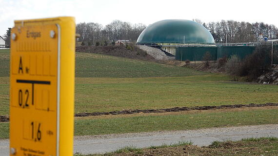 Eine grüne, kuppelartige Struktur im Hintergrund, umgeben von einem Feld und Bäumen. Im Vordergrund ist ein gelbes Messgerät mit Angaben zur Energie sichtbar.