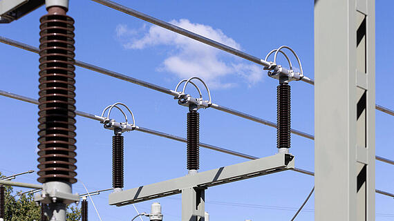 Eine Nahaufnahme von elektrischen Komponenten in einem Umspannwerk, einschließlich Isolatoren und Spannungsleitungen, vor einem blauen Himmel mit wenigen Wolken. Die Struktur zeigt solide metallische Teile, es sind keine Menschen sichtbar.