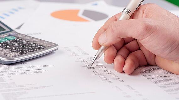 Eine Hand hält einen Stift und markiert Daten auf einem Finanzdokument. Neben dem Dokument liegt ein Taschenrechner und im Hintergrund sind Diagramme zu sehen.