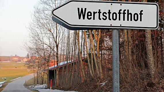 Ein Schild mit dem Begriff Wertstoffhof an einer Straße. Im Hintergrund ist ein Dorf zu sehen.