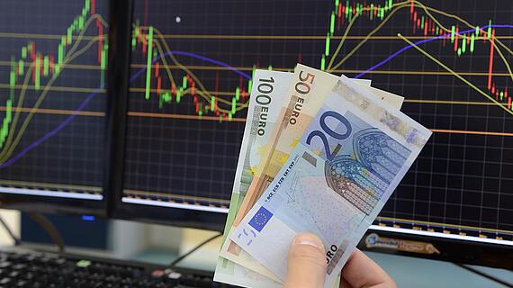 Jemand hält drei Geldscheine vor zwei Bildschirmen, die Kurstabellen anzeigen.