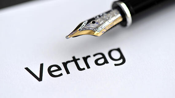 Eine Füllfederhalter-Spitze neben dem Wort "Vertrag", das auf einem weißen Blatt Papier gedruckt ist. Der Fokus liegt auf der Feder und dem Text.