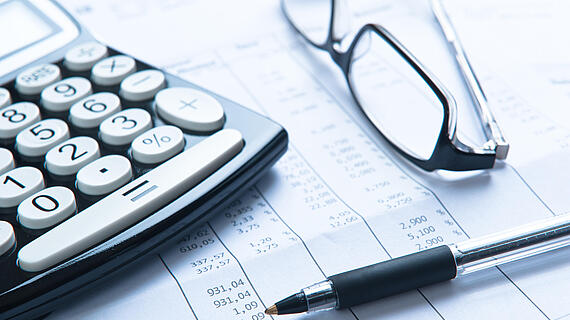 Ein Taschenrechner, eine Brille und ein schwarzer Kugelschreiber liegen auf einem Dokument mit Finanzdaten und Zahlen.