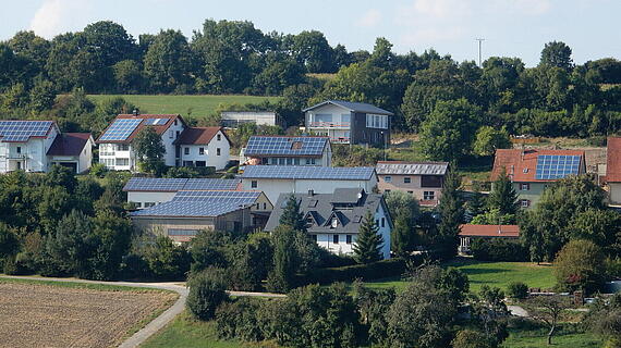 Landschaft mit mehreren Wohnhäusern, die Solarpanels auf den Dächern installiert haben. Im Hintergrund sind Bäume und ein hügeliges Feld zu sehen, unter einem klaren Himmel.