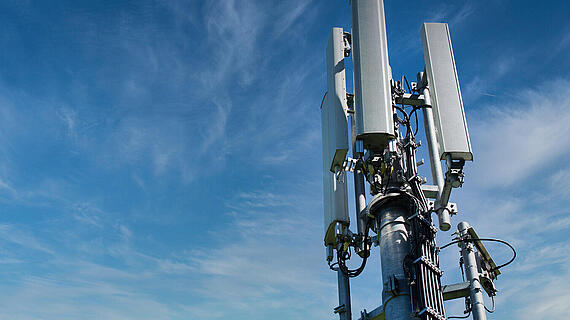Ein Mobilfunkmast mit mehrere Antennen vor einem blauen Himmel mit wenigen Wolken. Der Mast ist aus Metall und in der unteren Hälfte sichtbar, während die Antennen nach oben ragen.